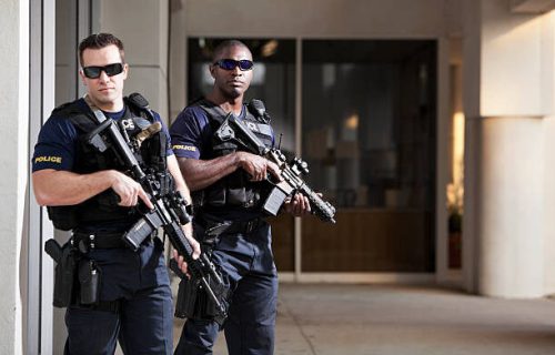 Multi-ethnic police officers (20s) wearing bulletproof vests, holding rifles.  Main focus on African American man.